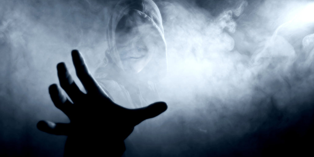 Pessoa com capuz surgindo de lugar escuro com fumaça, detalhe para a mão dela que vem em direção a quem olha a imagem