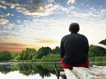 Homem está sentado em piso de concreto e admirando a paisagem com lago, floresta e céu azul com algumas nuvens